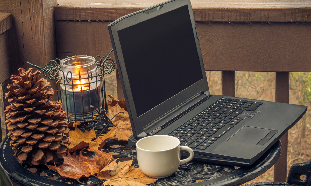 A black laptop sits on a small outdoor table on a wooden deck, surrounded by autumn leaves, a lit candle in a wire holder, a pinecone, and a white ceramic mug.